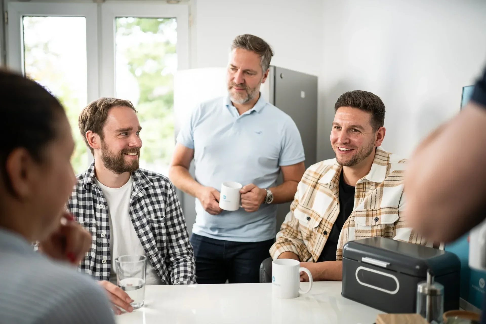 Vier Personen sitzen lachend um einen Tisch, ein weiterer steht mit einer Tasse daneben.